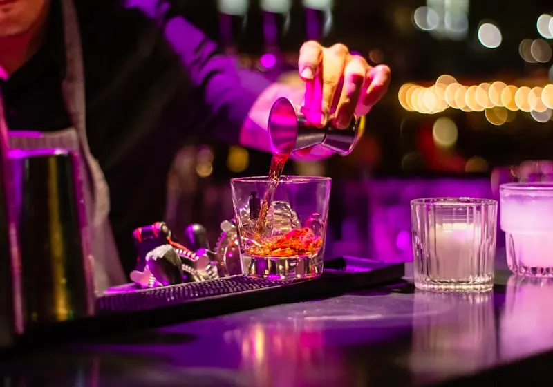 Barman versant un cocktail ambré dans un verre, ambiance lumineuse violette de bar à cocktails
