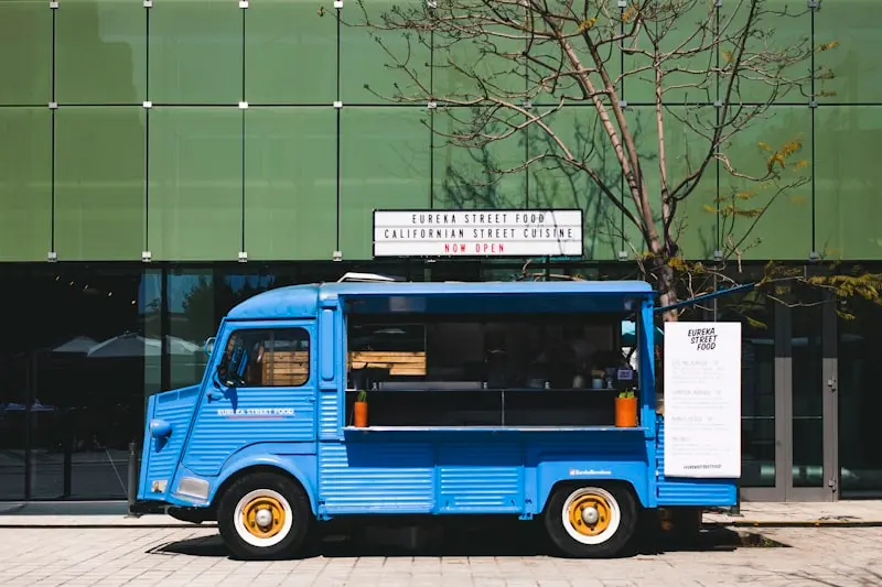 Food truck bleu vintage garé devant une façade vitrée avec enseigne lumineuse "Now Open"